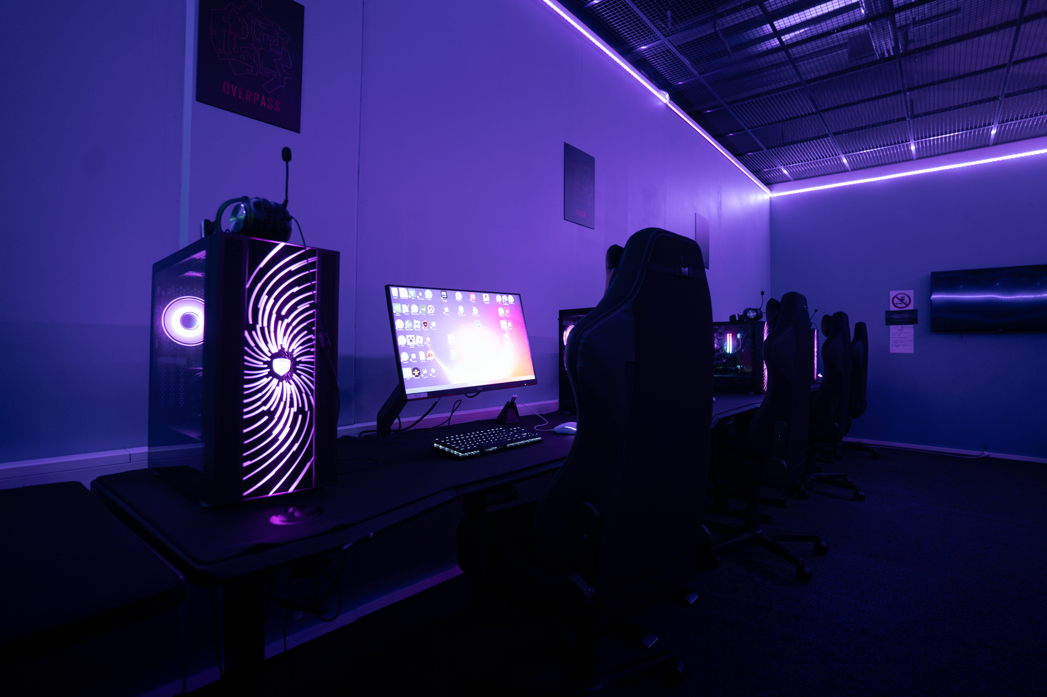 Kuva violetiksi RGB-valaistuksella värjätystä PC-huoneesta, kuvassa näkyy penkkirivi, yksi tietokone ja pelinäyttö.